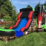 Colorful inflatable slide with a blue sliding area, red and green accents, and a brick-patterned base, set up on a grassy lawn with trees and bushes—perfect for NY party rentals Long Island events!.