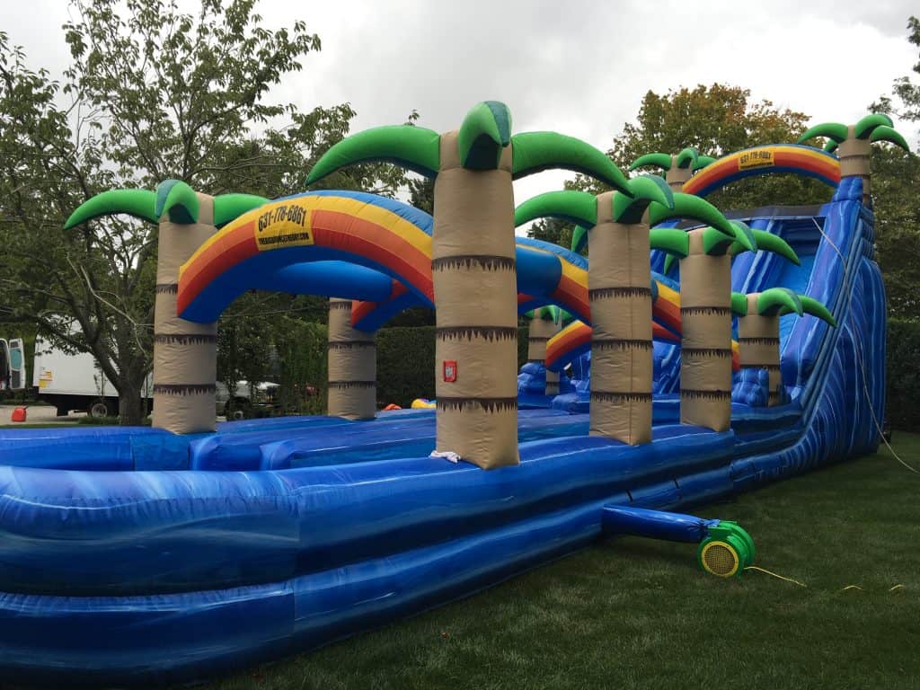 A large blue inflatable water slide with palm tree decorations and rainbow arches, perfect for party rentals Long Island, NY, set up on a grassy lawn with trees and a cloudy sky in the background.
