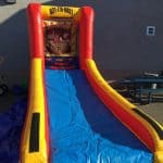 An inflatable slide with red, yellow, and blue colors is set up outdoors on a tarp next to a picnic table. Perfect for party rentals Long Island, NY, the slide features a sign at the top with a phone number and website.