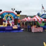 Two colorful bounce houses, including a Mickey Mouse-themed one, are set up beside red-and-white striped tents at an outdoor event in a Long Island parking lot. For fun gatherings like this, choose party rentals Long Island, NY locals love!.