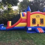 A colorful inflatable bounce house Long Island, NY with a slide sits on a grassy lawn, surrounded by trees. The red, yellow, and blue bounce house features pointed towers and mesh windows—perfect for party rentals Long Island!.