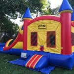 A large, colorful bounce house Long Island with towers and a slide sits on a grassy lawn, surrounded by trees. The red, yellow, and blue inflatable features mesh windows and a small entrance in front—perfect for party rentals Long Island, NY.