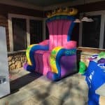A large, colorful inflatable throne with a crown-shaped backrest sits on a patio alongside a table with props and a white photo booth kiosk—perfect for party rentals Long Island. Nearby, there’s a building with stone and wood siding.