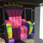 A large inflatable chair resembling a royal throne—perfect for party rentals Long Island—mostly pink with purple, yellow, and green accents, sits on a porch under a roof. Sunlight casts shadows across the chair.