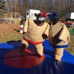 Two people wearing inflatable sumo wrestling suits face each other on a blue mat outdoors. The scene, set in a park with fallen leaves and party rentals Long Island equipment in the background, captures a fun-filled moment.