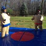 Two people in inflatable sumo wrestler suits face off on a blue mat outdoors, surrounded by trees and mini-golf obstacles—perfect for any party rentals Long Island event, while another person watches the fun in the background.