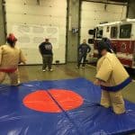 Two people in inflatable sumo suits face off on a blue mat with a red circle, as three others—including a firefighter—watch near a fire truck in a garage, showcasing fun party rentals Long Island can offer for events.