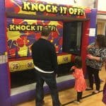 Two adults and two children stand in front of the "Knock It Off!" inflatable game, often featured at NY party rentals Long Island events. Inside a gym, they get ready to throw balls at colorful targets with point values.