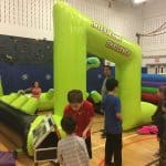 Children play and wait in line around a large green inflatable obstacle course labeled "Interactive Challenge" inside a gymnasium. Adults supervise nearby, and a climbing wall is visible in the background—perfect for party rentals Long Island, NY.