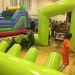 Children play an interactive game on a green bounce house Long Island obstacle course indoors, pressing light-up buttons. A digital scoreboard displays 27 and 29 as other kids and adults watch in the background.