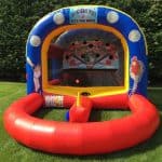 An inflatable baseball-themed game setup on grass, featuring a blue and yellow arch with baseball graphics, two cartoon players, a red front barrier, and a ball aimed at holes—perfect for party rentals Long Island events.