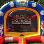 An inflatable baseball game setup with cartoon players and pitching targets, perfect for party rentals Long Island, is displayed outdoors on grass. A sign reads “The Big Boom Theory” with a phone number, surrounded by green bushes.