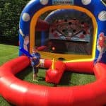 A young boy stands with a plastic bat in front of a colorful bounce house Long Island baseball game setup on grass, ready to hit a ball toward targets inside the inflatable structure decorated with baseball players.