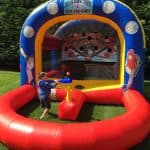 A young boy plays a baseball game, swinging a bat at a floating ball in front of an inflatable baseball-themed party rentals Long Island game structure on a grassy lawn.
