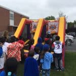 Children stand in line outdoors, waiting to play on an inflatable basketball game from party rentals Long Island. The weather is cloudy, and the activity is next to a brick building and a chain-link fence with parked cars in the background.