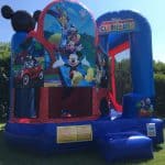 A colorful Mickey Mouse Clubhouse-themed bounce house sits on grass in NY, decorated with large images of Mickey, Minnie, and friends. Perfect for party rentals Long Island, it features a blue and red design with steps. Trees and a blue sky are in the background.