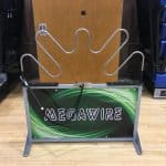 A metal "Megawire" game with a curvy wire path, a handle on a cord, and a green and black sign, set on a gym floor between blue bleachers and a wooden door—perfect for NY party rentals Long Island events.
