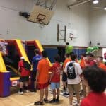 Children and staff gather in a gymnasium, forming a line in front of a colorful bounce house Long Island obstacle course. Most children wear orange shirts, and basketball hoops are visible overhead.