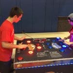 Two people in red shirts play a fast-paced arcade game with colorful, light-up buttons at a gymnasium. This NY event, featuring fun party rentals Long Island style, brings excitement and competition to any gathering.