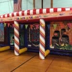 Inflatable carnival games booth indoors with colorful striped columns, bunting, and three different game sections; great for party rentals Long Island, NY. A U.S. flag hangs on the wall in the background.