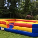 A large, colorful bounce house Long Island with red, yellow, and blue sections sits on a grassy lawn surrounded by trees and bushes. A person stands nearby, looking at the inflatable.