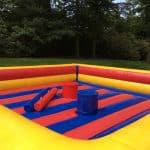 Inflatable jousting arena with red, yellow, and blue stripes, featuring two padded platforms and padded jousting sticks. Perfect for party rentals Long Island, this arena sits on grass surrounded by trees and greenery.