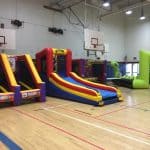 Inflatable obstacle courses and slides in bright colors are set up inside a gymnasium with basketball hoops, wooden floors, and high ceilings. Perfect for party rentals Long Island, these inflatables include slides and game stations for endless fun.