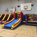 Three colorful inflatable games, available through party rentals Long Island, are set up in a gymnasium, including a slide, a basketball game, and a "Knock It Off!" target game, with basketball hoops and wooden flooring visible in the background.
