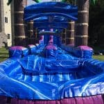 A close-up view of an inflatable blue and purple water slide, set up outdoors on a grassy lawn near a beige house and surrounded by trees—perfect for NY parties and available from bounce house Long Island party rentals.