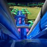 View from the top of a large blue inflatable water slide with palm tree decorations, looking down to a grassy yard—perfect for party rentals Long Island or NY celebrations!.