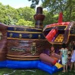 Three people stand near a large inflatable bounce house Long Island shaped like a pirate ship, set up outdoors on green grass with trees behind. The colorful party rentals NY ship features cannons and a slide.