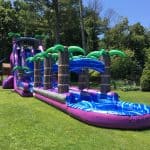 A large inflatable water slide with blue and purple sections and palm tree decorations, available from party rentals Long Island, NY, is set up on a grassy lawn surrounded by tall trees and lush greenery.