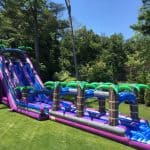 A large, colorful inflatable water slide with palm tree decorations stands on a grassy lawn, leading to a slip-and-slide and splash pool—perfect for party rentals Long Island events, surrounded by lush trees and greenery.