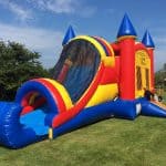 A colorful inflatable bounce house Long Island with blue, red, and yellow sections sits on green grass under a blue sky, featuring a slide and turrets resembling a castle—perfect for NY party rentals Long Island events.