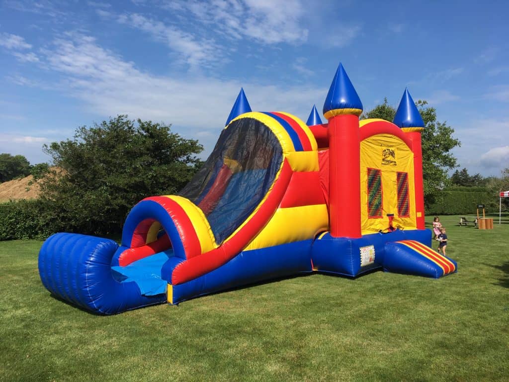 A large, colorful inflatable bounce house Long Island with a slide sits on a grassy lawn under a partly cloudy NY sky; a child stands nearby.