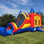 A large, colorful inflatable bounce house Long Island with a slide sits on a grassy lawn under a partly cloudy NY sky; a child stands nearby.