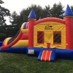 A colorful inflatable bounce house designed like a castle, perfect for party rentals Long Island, sits on a grassy lawn with trees in the background. The structure features vibrant red, yellow, and blue colors along with a fun slide.