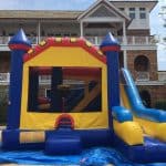 A colorful inflatable bounce house Long Island with a slide sits on a blue tarp in front of a large building with white railings and brown siding. The bounce house is mainly yellow, blue, and red—perfect for NY party rentals Long Island events.