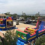 Two colorful inflatable bounce houses and obstacle courses from party rentals Long Island are set up outdoors near a parking lot, with a few people nearby and trees and buildings in the background. The area looks festive and playful.