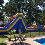 A large, colorful inflatable water slide from party rentals Long Island is set up on a grassy backyard near a swimming pool, with patio chairs and potted plants in the foreground and tall trees in the background.