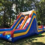 A large inflatable slide with blue, yellow, and red sections stands on a grassy yard in NY, surrounded by trees and a wooden fence—perfect for bounce house Long Island party rentals.