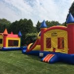 Two colorful inflatable bounce houses sit on a green lawn in NY; one has a slide attached, both are red, yellow, and blue—perfect for party rentals Long Island. They're surrounded by trees under a partly cloudy sky.