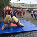 Two people in inflatable sumo suits wrestle on blue mats outdoors in NY, cheered on by onlookers near tents and a large building. Some spectators wear red or white shirts at this lively party rentals Long Island event.
