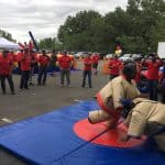 Two people in inflatable sumo suits wrestle on a blue mat outdoors, surrounded by a cheering crowd in matching red shirts. The fun scene, perfect for party rentals Long Island, NY, features trees, tents, and parked cars in the background.