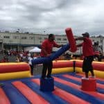 Two people in red shirts balance on pedestals in a bounce house Long Island arena, jousting with padded batons, while a crowd watches at an outdoor NY event near a large building and trucks.