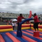 Two men in red shirts balance on inflatable pedestals and playfully joust with padded sticks in a colorful bounce house Long Island arena at an outdoor event, surrounded by party rentals and trucks in the background.