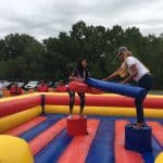 Two women stand on red pedestals in an inflatable jousting arena from a party rentals Long Island company, each holding padded jousting sticks as people watch in the background at this lively outdoor NY event.