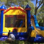 Three people are standing and crouching near a colorful inflatable bounce house with a slide in a sunny, tree-filled NY backyard—perfect for those searching for bounce house Long Island or party rentals Long Island.