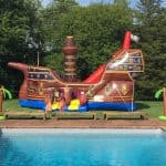 An inflatable pirate ship slide, part of our party rentals Long Island collection, is set up on grass behind a swimming pool. Two children stand at the entrance, with palm tree decorations on either side under a clear blue NY sky.