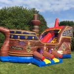 An inflatable pirate ship bounce house Long Island, complete with cannons and a slide, sits on green grass among trees under a partly cloudy sky—a perfect choice from party rentals Long Island for your next NY event.